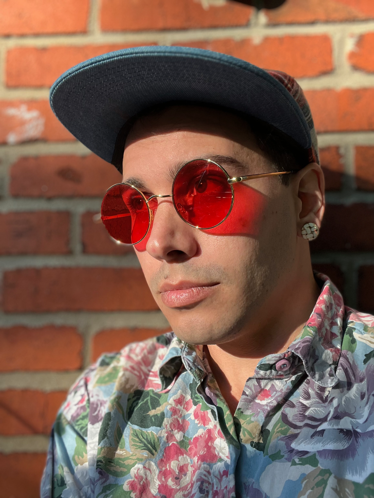 BeWyrd Headshot. Poet stares into sun with red sunglasses and a brick background
